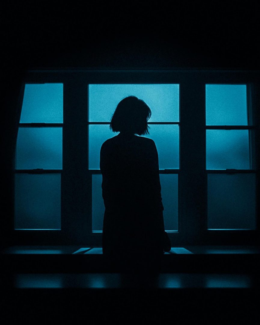 a woman stands in a blue toned dimly lit room. Her back is to the camera. She quietly contemplates while looking slightly beond frame